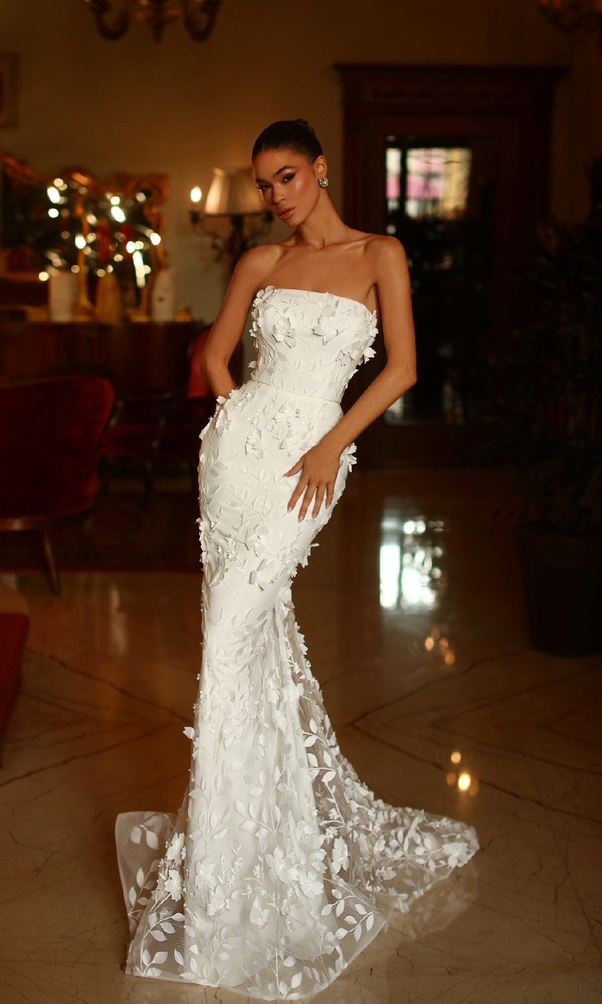 A woman stands indoors on a polished marble floor in an elegant, dimly lit room wearing the Puff-Sleeve White Formal Gown: Tina Holly TW121W, a form-fitting gown with 3D floral appliqués.
